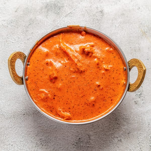 Overhead view of a metal bowl filled with vibrant orange Nepalese butter chicken on a light background.