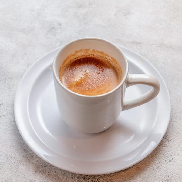 A white mug filled with dark coffee and foam, sitting on a white saucer.