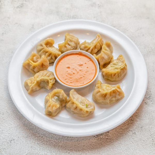A white plate holds a dozen steamed Nepalese momo dumplings surrounding a bowl of orange dipping sauce.